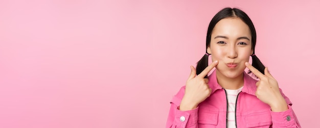 Free photo close up portrait of young asian girl showing her dimples poking cheeks silly and making funny faces standing over pink background