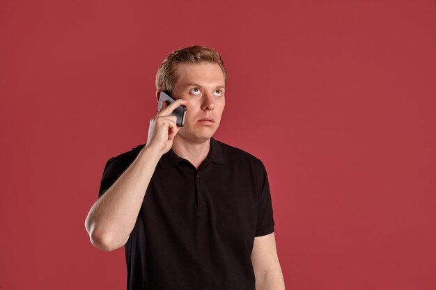 Close-up portrait of a young athletic ginger man in a stylish black t-shirt looking thoughtful and talking by a smartphone while posing on pink studio background. Human facial expressions. Sincere emo