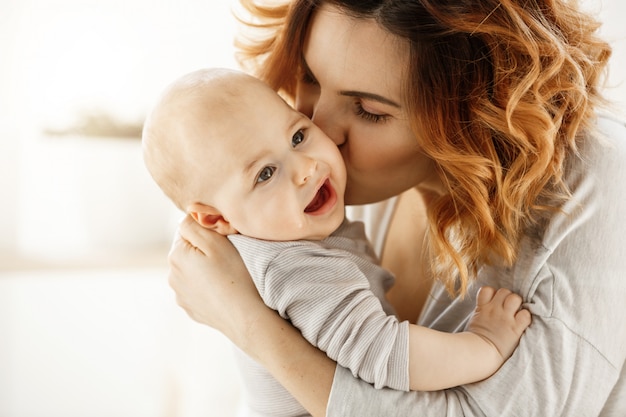 Free photo close up portrait of young attractive mother kissing his crying baby. kid looking aside with frightened face expression. family concept.