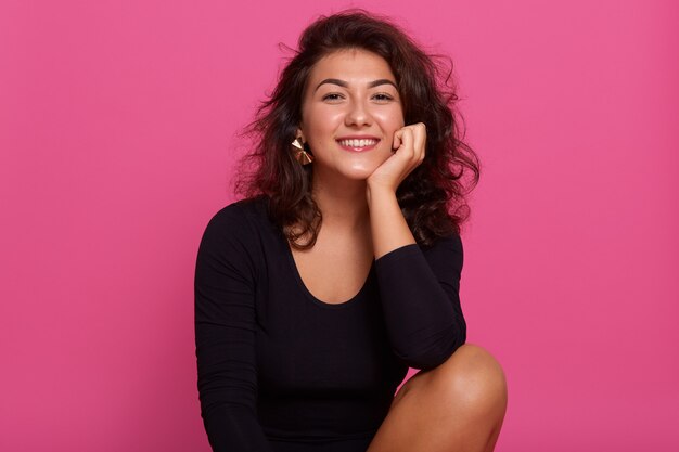 Close up portrait of young cheerful beautiful girl with dark wavy hair dressed black shirt, sitting smiling with teeth against pink wall