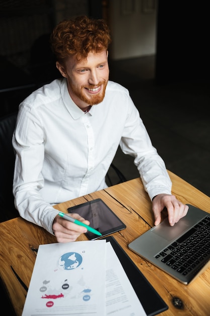 Free photo close-up portrait of young cheerful redhead curly business man in white shirt working on laptop, looking aside