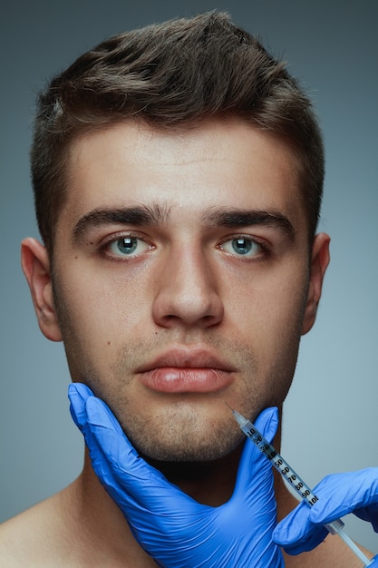 Close-up portrait of young man isolated on grey studio background. Filling surgery procedure. Concept of men's health and beauty, cosmetology, self-care, body and skin care. Anti-aging.