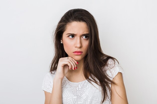 Close-up portrait of young thinking woman in white blouse, frowning, holding hand at her face, looking aside, serious face expression, having a problem isolated