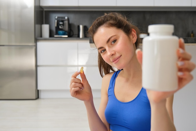 Free photo close up portrait of young woman fitness instructor showing bottle of vitamins taking buds