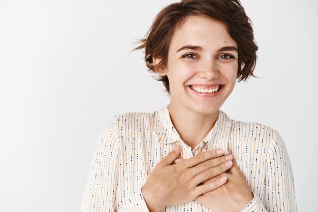 Close up of pretty girl with natural makeup hold hands on heart and smile with grateful face saying thank you standing over white background