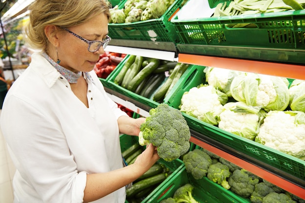 Free photo close-up profile portrait of good-looking middle-aged caucasian woman vegetarian in casual clothes picking-up and choosing the freshest vegetables and broccoli at grocery store. people and shopping