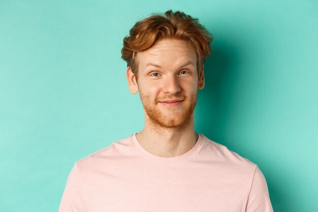 Close up of redhead bearded man looking pleased, nod in approval and smiling, standing in pink t-shirt against turquoise background.