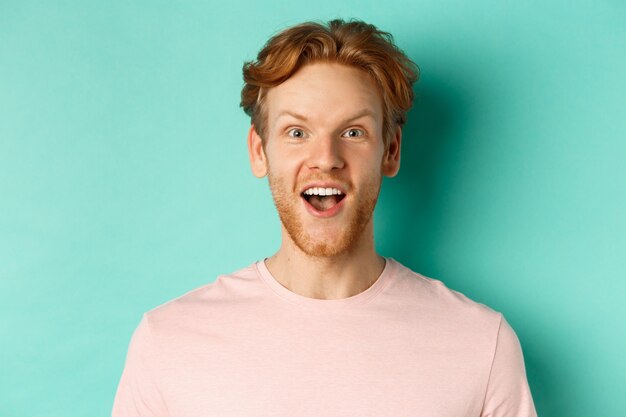 Close up of redhead bearded man raising eyebrows and looking surprised at camera, checking out promotion, standing over mint background.