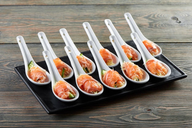 Free photo close-up of salmon appetizers set: black board served with white spoons,including smoke salmon rolls with cucumber and cheese. delicious dish for light alcohol drinks, buffet or catering.