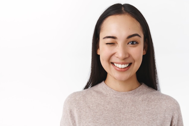 Free photo close-up sassy, enthusiastic happy smiling asian woman with long dark hair, natural nude make-up, having great day, send positive vibes, winking to cheer friend up and say everything under control