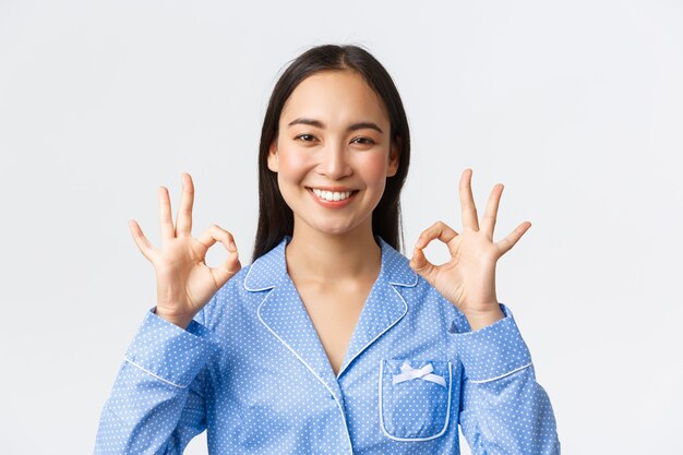 Close-up of satisfied happy asian woman in blue pajama rate nice work, recommend perfect product or service quality, guarantee something as show okay gesture pleased, white background