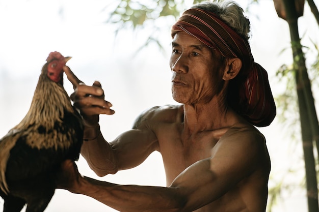 Free photo close up, selective focus face of asian senior man shirtless and turban with loincloth, sitting and touch the mouth of a gamecock. he teases the beautiful gamecock with love. copy space