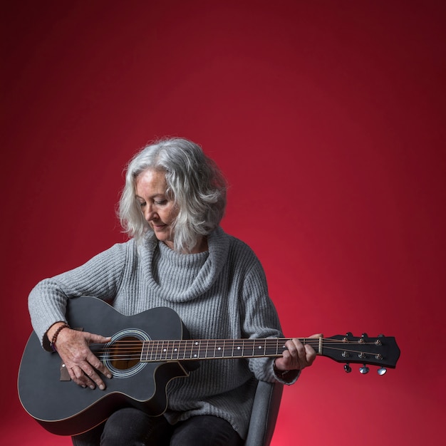 Close-up of senior woman sitting in chair playing the guitar against red background