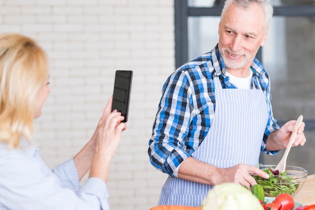 Free photo close-up of senior woman taking photo of her husband preparing the salad in the bowl on mobile phone