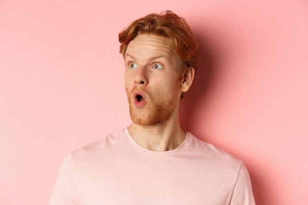 Close up of shocked redhead man with beard, saying wow, looking left with amazed face, standing over pink background