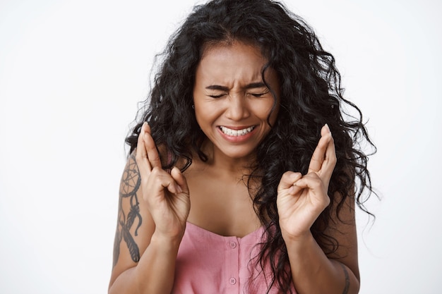 Close-up shot anxious and worried curly-haired female with tattoos, squinting, clench teeth nervously, cross fingers good luck, anticipating important thing, praying standing white wall