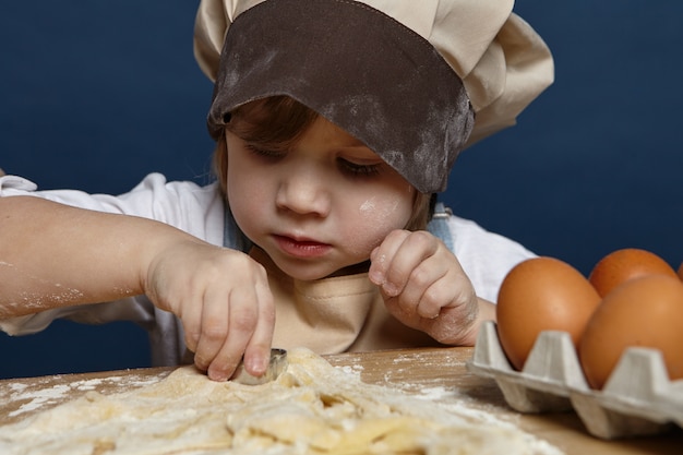 Close up shot of charming cute little girl wearing big chef cap making cookies at the kitchen table, using bakery moulds, having focused concentrated expression. Children, cooking and baking concept