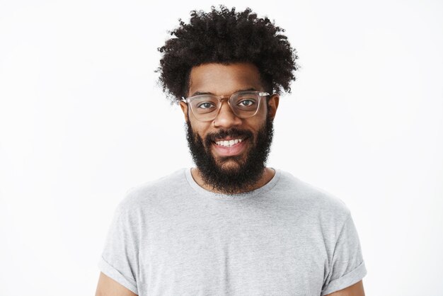 Close-up shot of charming pleasant and delighted dark-skinned bearded male customer in glasses with afro hairstyle in grey casual t-shirt, smiling joyfully as having great mood on gray.