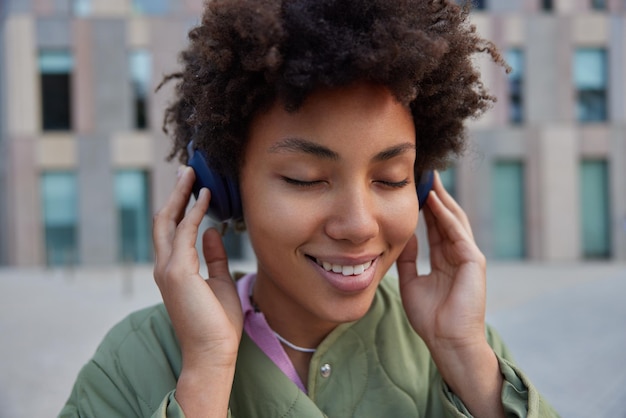 Free Photo close up shot of cheerful curly haired woman closes eyes from pleasure while listens favorite audio song via headphones wears jacket