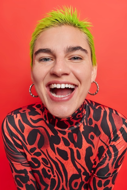 Free photo close up shot of cheerful freckled teenage girl smiles broadly shows white teeth has trendy ideas gathers with friends of same age have fun poses against red wall