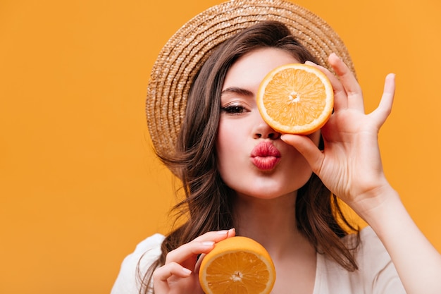 Free photo close-up shot of cute girl in straw hat covering eye with orange and blowing kiss.