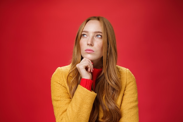 Close-up shot of determined and focused creative thoughtful redhead female looking at upper left corner touching chin, thinking, making choice or remembering information over red background.