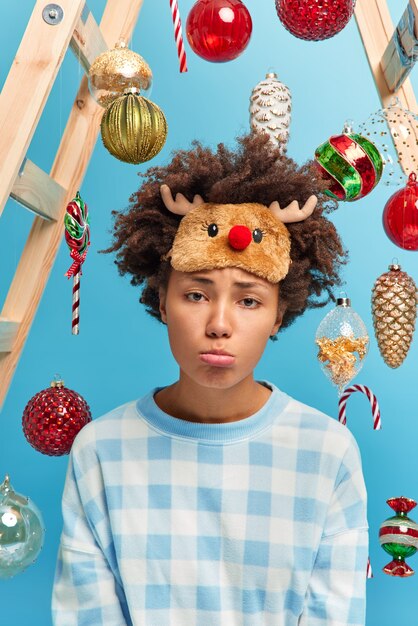 Close up shot of displeased Afro American woman looks with gloomy face expression at camera sad to be ill during winter holidays wears warm sleep mask and pajama decorates house with New Year trees