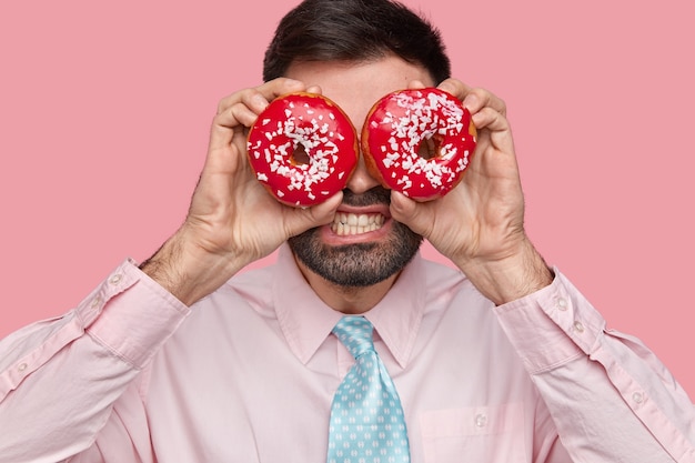 Free photo close up shot of displeased bearded man clenches teeth, covers eyes with doughnuts, dressed formally, stands over pink wall