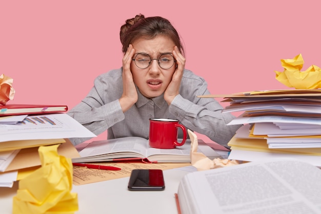 Free photo close up shot of dissatisfied woman keeps hands on temples, frowns face, has fatigue expression, wears round spectacles