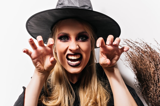 Close-up shot of evil witch with gray eyes. Indoor photo of scary female vampire in black hat.