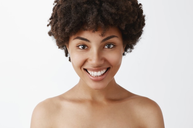 Close-up shot of flirty and feminine confident african american woman with broad bright smile posing naked being satisfied with great skin condition after cosmetological treatment