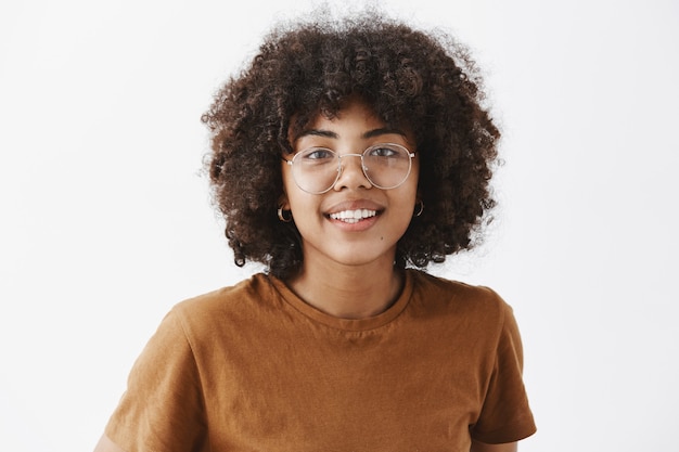 Close-up shot of friendly good-looking african american woman in transparent glasses and brown t-shirt smiling with joyful pleasant smile being satisfied with how things going over gray wall