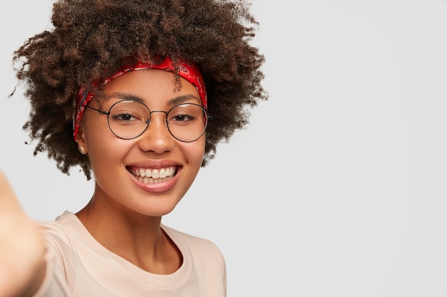 Close up shot glad feminine girl has toothy smile, Afro hairstyle, beautiful clean skin, wears transparent glasses, stretches hand as holds unrecognizable device, makes selfie over white wall