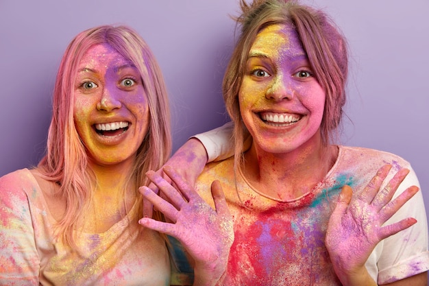 Free photo close up shot of glad smiling women being in high spirit, smile broadly, show palms, have dirty faces and palms with holi colors, have fun indoor, isolated over purple wall. celebration