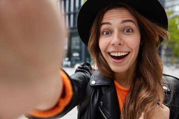 Close up shot of glad woman wears black hat, smiles broadly, being in high spirit, has fun during walk outside