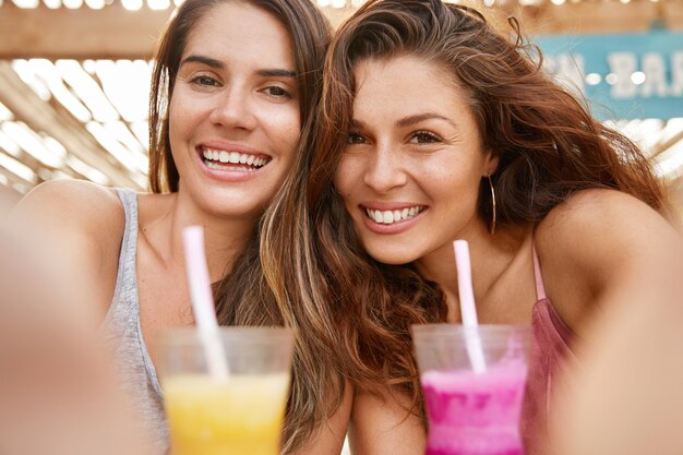 Close up shot of good looking European women have fun, celebrate something, drink summer cocktails, make selfie, share photos in social networks.