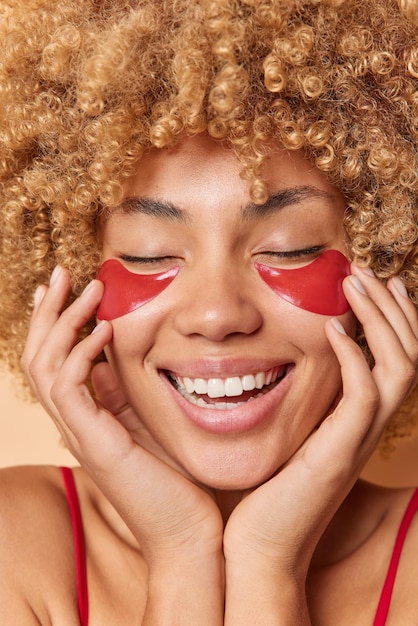 Free photo close up shot of happy curly haired woman applies red hydrogel patches under eyes for skin treatment touches face gently smiles broadly shows white teeth stands very happy indoor beauty concept