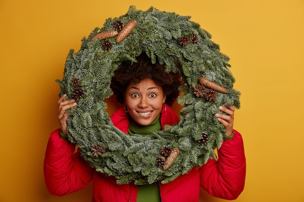 Free photo close up shot of happy dark skinned woman looks through round spruce wreath, dressed in red jacket, makes shot against yellow wall, happy her favourite winter holidays come soon.