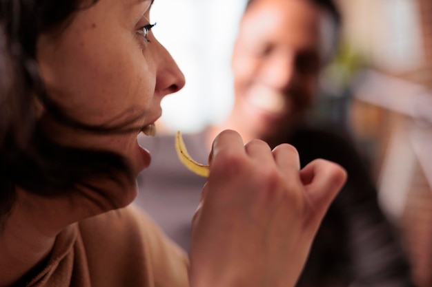 Close up shot of happy woman eating chips while sitting at home with multiethnic friends. Cheerful person enjoying snacks while sitting at table in living room with diverse people.