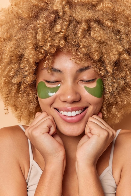 Close up shot of happy woman with healthy complexion curly bushy hair smiles toothily keeps eyes closed applies green hydrogel patches for skin refreshment. Beauty time and face care concept