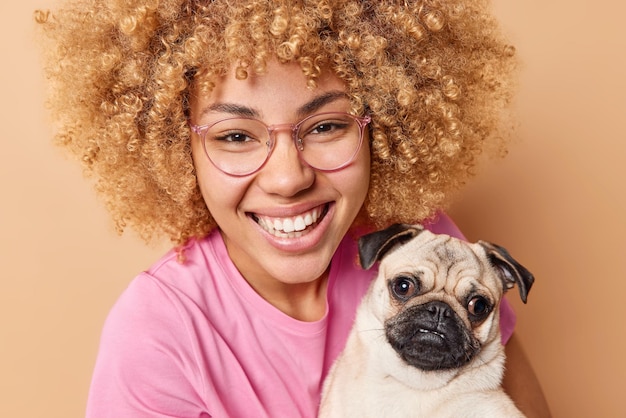 Free Photo close up shot of happy young woman with curly hair smiles gladfully enjoys company of pet poses with pug dog dressed casually isolated over beige background people and domestic animals concept