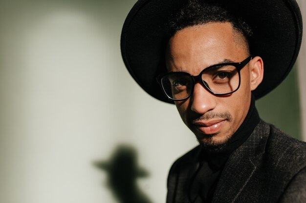 Close-up shot of interested guy in glasses posing in dark room. Portrait of black male model in trendy hat isolated on gray wall.