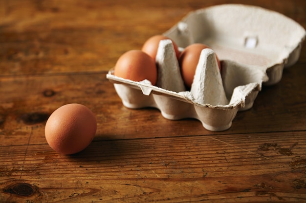 Close up shot of an open recyclable egg carton with 3 eggs inside and one egg next to it on a brown grainy wooden table