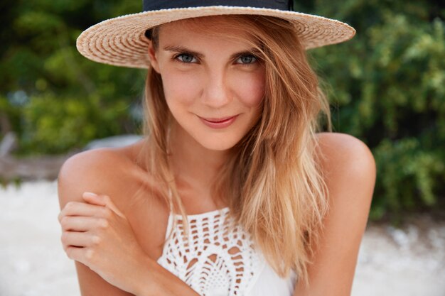 Close up shot of pleasant looking with long hair, has pure healthy skin, wears straw hat, poses outdoor, recreate in paradise place with lover, has pleased expression. Summer time and rest concept