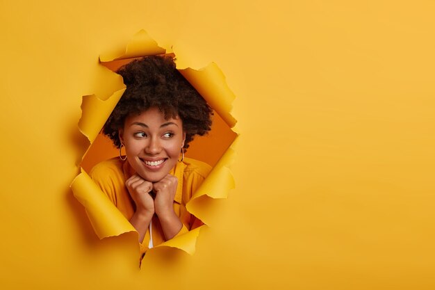 Close up shot of pretty cheerful woman holds both hands under chin, expresses friendly joyful attitude, stands upbeat, wears yellow clothing, looks away, poses in ripped background