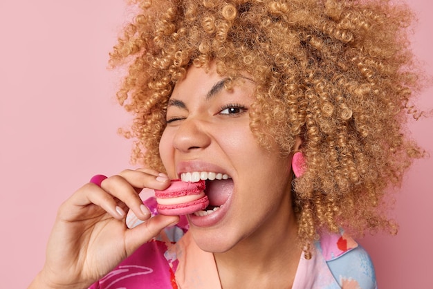 Free photo close up shot of pretty curly haired woman eats delicious french macaroon enjoys dessert bakery sweet yummy food keeps mouth widely opened poses against pink background unhealthy eating concept