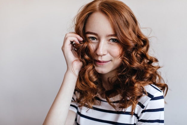 Close-up shot of refined ginger woman posing with shy smile. Indoor photo of graceful white lady expressing positive emotions.