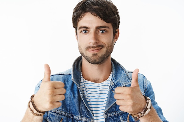 Free photo close-up shot of satisfied and happy young attractive male with blue eyes and bristle in denim jacket raising thumbs up and smiling cheerfully