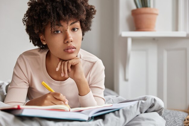 Close up shot of serious dark skinned female college student does home assigment in bed, writes in notebook with pen, keeps hand under chin