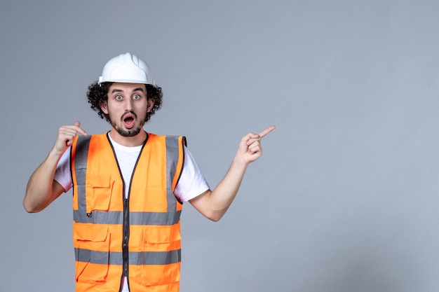 Close up shot of shocked male constructor in warning vest wearing safety helmet and pointing something on the left side over gray wave wall